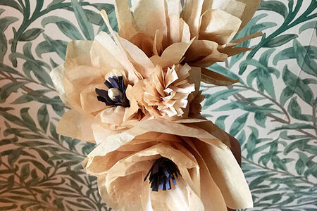 Two kraft paper flowers with dark paper centres. Green and white leaves and stem print wallpaper serves as the background.