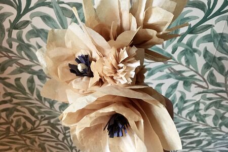 Two kraft paper flowers with dark paper centres. Green and white leaves and stem print wallpaper serves as the backgroun